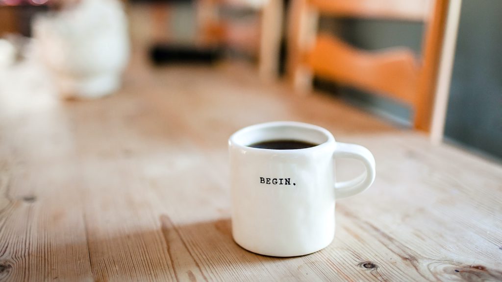 A white coffee mug with the word "begin" on it sits on a wooden table where you can sit and prayer these 5 powerful morning prayers.