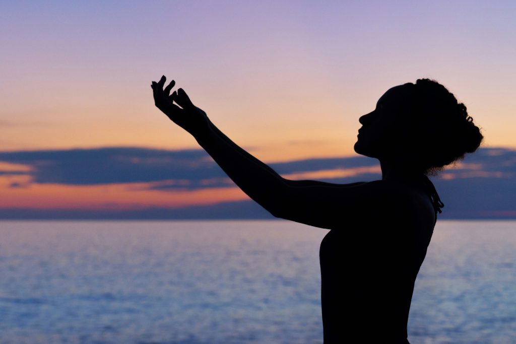 A woman in silhouette stands on the shore at sunset lifting her arms to God in thanks.